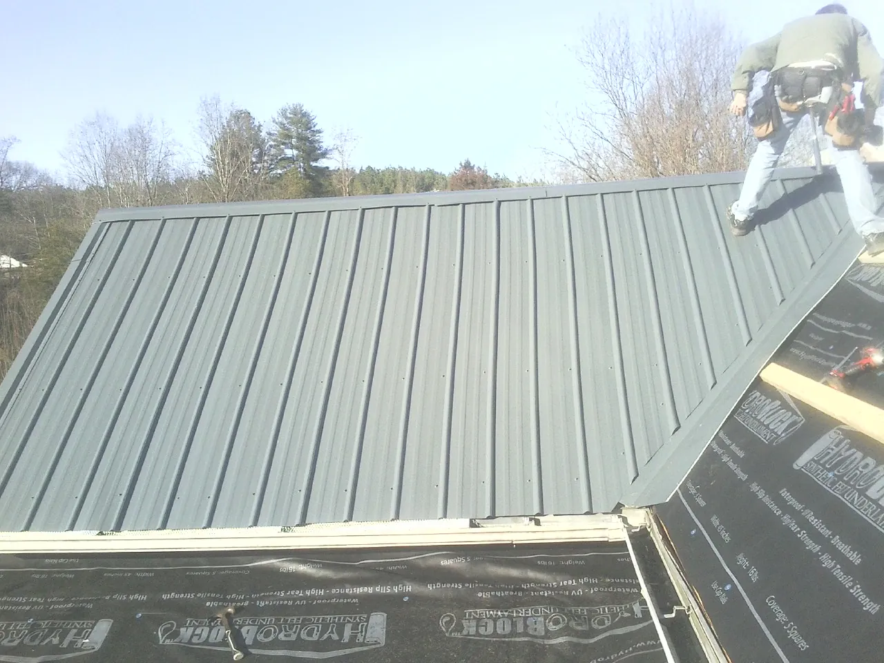 Crew installing dark green standing seam panels over synthetic underlayment in Benton